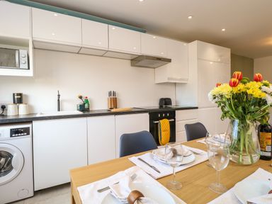 A kitchen with white cabinets and black countertop next to a wooden dining table set with plates glasses and a vase of flowers at Seaside House in Gorleston-On-Sea