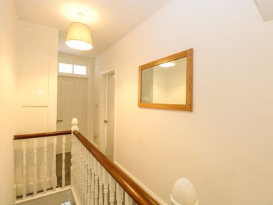 A hallway with white walls a wooden handrail white balusters a door a mirror and a ceiling light at Seaside House in Gorleston-On-Sea