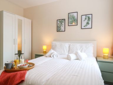 A bedroom with a bed with white bedding and rolled towels bedside tables with lamps a wardrobe and framed leaf prints on the wall at Seaside House in Gorleston-On-Sea