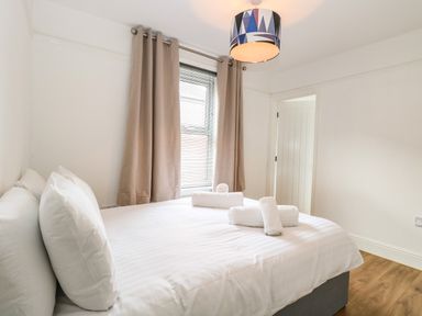A bedroom with a double bed with white bedding and pillows next to a window with beige curtains at Seaside House in Gorleston-On-Sea