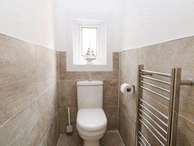 A bathroom with a toilet a toilet brush a window with a small sailboat decoration a toilet paper holder and a metal towel rack at Seaside House in Gorleston-On-Sea