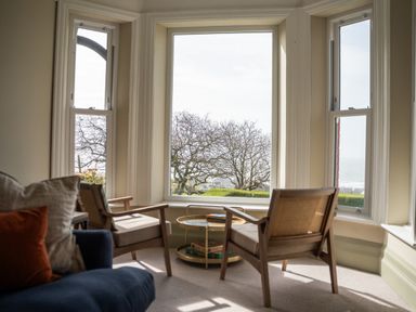 A sitting area with two wooden armchairs and a round side table near large windows overlooking trees and a distant view at Haulfryn in Barmouth