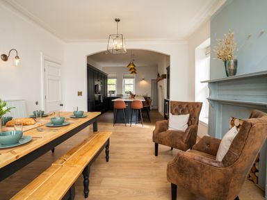 A dining area with wooden table and benches next to a seating area with two brown chairs and a blue fireplace at Haulfryn in Barmouth