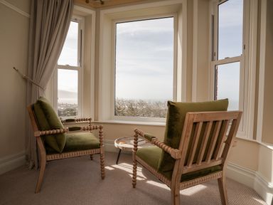 A corner with two wooden armchairs with green cushions and a round side table by large windows at Haulfryn in Barmouth