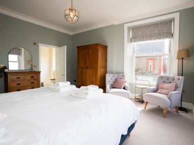A bedroom with a bed, two armchairs with cushions, a wooden wardrobe, a chest of drawers with a mirror, and a floor lamp at Haulfryn in Barmouth