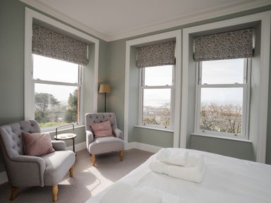 A bedroom with two gray armchairs with pillows a floor lamp and three windows with patterned blinds at Haulfryn in Barmouth