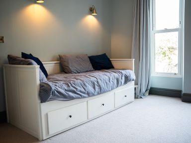 A bedroom with a white daybed with gray bedding and blue pillows under wall lights next to a window with curtains at Haulfryn in Barmouth
