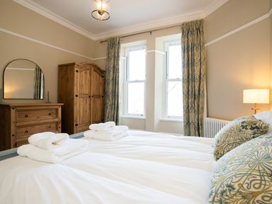A bedroom with a bed with pillows and folded towels two windows wooden wardrobe dresser and a lamp at Haulfryn in Barmouth