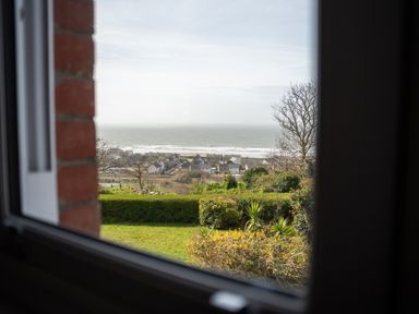 View through a window of a garden with bushes and trees overlooking houses and the sea at Haulfryn in Barmouth