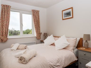 A bedroom with a double bed with pillows and rolled towels next to a window with patterned curtains at Mabel's View in East Allington Devon