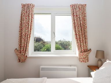 A bedroom window with patterned curtains a bedside table with a lamp and a white bed at Mabel's View in East Allington Devon