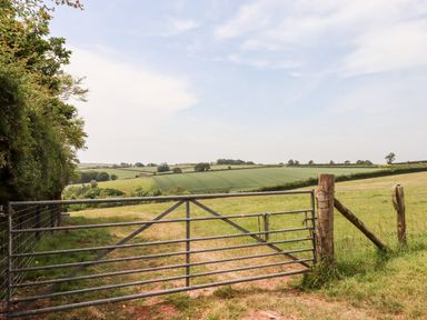 A metal gate opening to green fields with rolling hills and trees under a blue sky at Mabel's View in East Allington Devon
