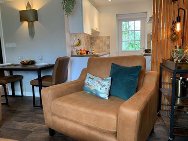 A seating area with a brown armchair and cushions next to a dining table and kitchen at Inny Brook Burrow in Davidstow near Camelford