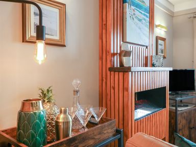 A wooden tray with a green vase a golden pineapple a metallic shaker a glass decanter and martini glasses next to a wooden fireplace and a TV in a living room at Inny Brook Burrow Davidstow near Camelford