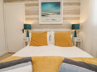 A bedroom with a bed with yellow pillows and a blanket two blue lamps on white bedside tables and a painting on a wooden wall at Inny Brook Burrow in Davidstow near Camelford