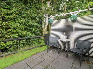 An outdoor patio with two black chairs a round metal table and a wooden slatted fence with hanging plants at Inny Brook Burrow in Davidstow near Camelford