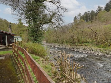 A river flowing beside a wooden fence with trees and a cabin nearby at Chestnut Timber Lodge in Keswick