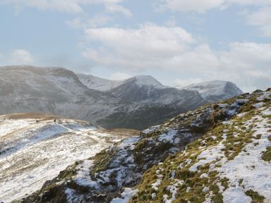 Snow partially covering grassy hills with mountain range in the background under a cloudy sky