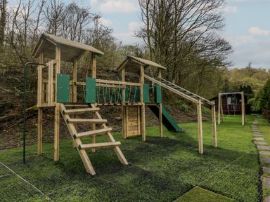 A wooden playground set with swings and climbing sections on grass near trees at Dogwood Timber Lodge in Keswick