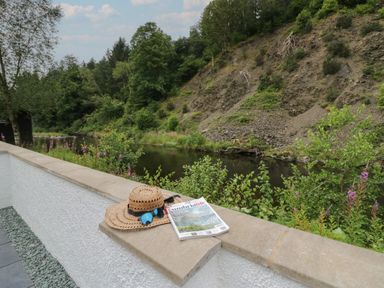 A magazine and a straw hat with sunglasses resting on a wall by a river with trees and a rocky hill in the background at Riverside Cottage No 4 in Keswick
