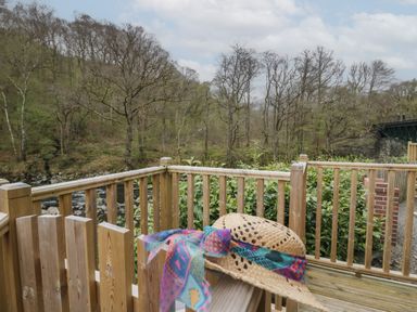 A wooden deck with a straw hat on the railing overlooking trees and a river at Pod No 6 Keswick