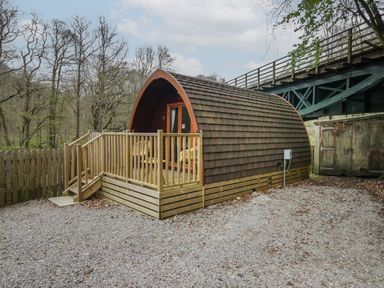 A small wooden pod with a deck and chairs next to a bridge in a wooded area at Pod No 28 in Keswick