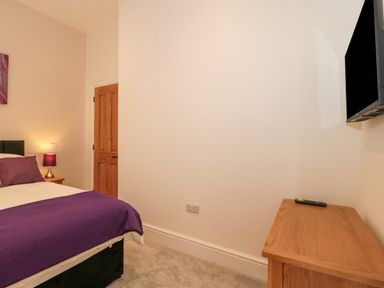 A bedroom with a bed with purple bedding a wooden door a wooden dresser with a remote and a wall-mounted television at The Nest in North Petherton