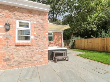 A stone exterior wall with a window outdoor hot tub with steps and a retractable awning on a paved patio with a wooden fence and trees in the background at The Nest in North Petherton