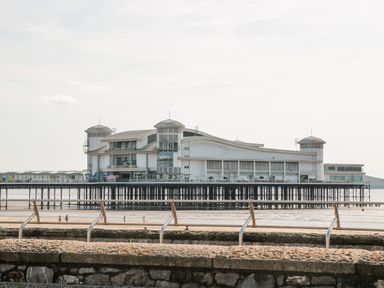 A pier building over water with people walking on the beach behind a stone wall and metal railing