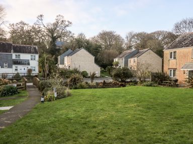 A grassy lawn with a small bridge and a pathway leading to several houses with trees in the background at Chellowdene Cottage in Falmouth