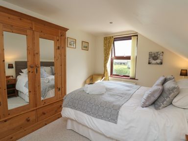 A bedroom with a bed with pillows and a gray patterned blanket a wooden wardrobe with mirrors a window with yellow curtains and two lamps at The Clove in Poyston Cross near Haverfordwest