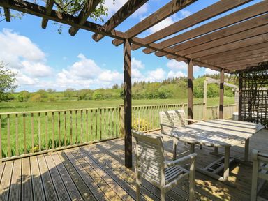 An outdoor wooden deck with a table and chairs under a pergola overlooking a green field at The Clove in Poyston Cross near Haverfordwest