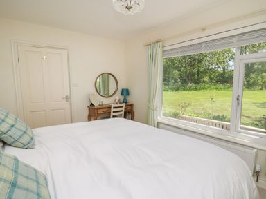 A bedroom with a bed with plaid pillows a wooden dressing table with a round mirror and a window showing a garden at Lavender Lodge in Haverfordwest