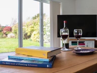 A wooden table with three books a bottle of wine two glasses of wine and a plate in a room with sliding glass doors at Little Chapter in Walberswick