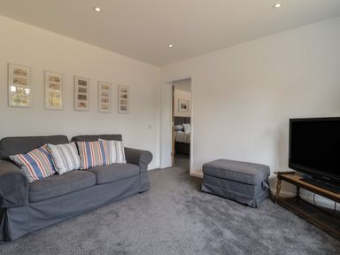 A living room with a gray sofa with striped pillows a gray ottoman a wooden TV stand with a television and framed pictures on the wall at Little Chapter in Walberswick