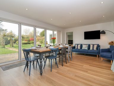 A dining area with a wooden table and six chairs set with glasses and dishes next to a seating area with blue sofas and a wall-mounted television at Little Chapter in Walberswick