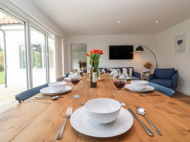 A dining table set with bowls plates cutlery glasses of wine and a bottle with flowers in a room with blue chairs a wall mounted tv and sliding glass doors at Little Chapter in Walberswick