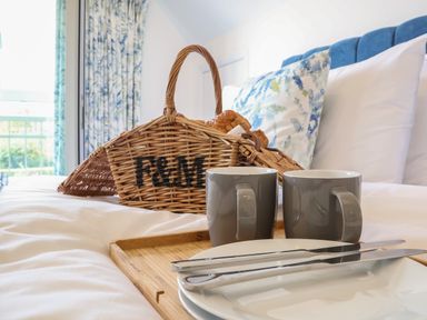 A bed with a wooden tray holding two grey mugs white plates and cutlery next to a wicker basket with bread at Little Chapter in Walberswick