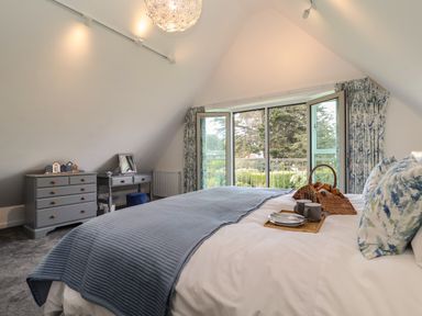A bedroom with a bed dressed in white and blue linens with a tray of cups and plates and wicker basket near a window at Little Chapter in Walberswick