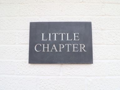 A black rectangular sign with the words Little Chapter mounted on a white textured wall at Little Chapter in Walberswick