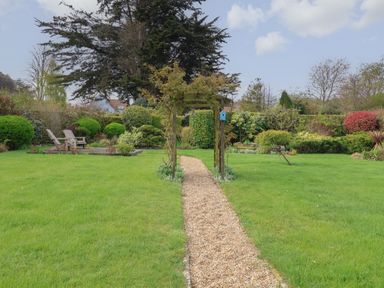 A garden with a gravel pathway leading to a wooden arch and two chairs on a raised wooden platform surrounded by bushes and trees at Little Chapter in Walberswick