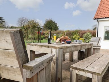 An outdoor wooden table and chairs with a basket of bread a bottle a glass of juice and cups in a garden at Little Chapter in Walberswick