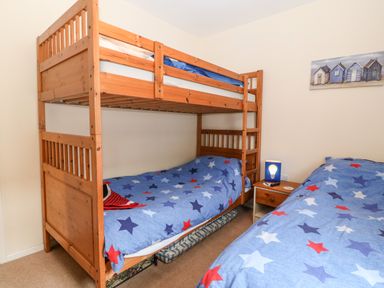 A bedroom with wooden bunk beds and a single bed with star patterned bedding a nightstand with a lamp and a picture of beach huts on the wall at Jacuma in Rhosneigr