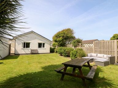 A backyard with a wooden picnic table and outdoor sofa near a white house with a bench at Jacuma in Rhosneigr