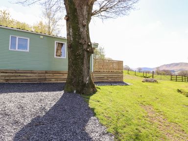 A large tree casting a shadow on gravel beside a green modular building with a wooden deck and a grassy field with a fence and distant hills at Whiteside in Lorton
