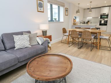 A living area with a grey sofa, a wooden coffee table, a dining table with chairs, and a kitchen in the background at Sebastian's View in Falmouth