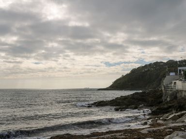 A rocky shoreline with waves and cloudy sky near a wooded hill and concrete structures at Sebastians View in Falmouth