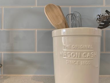 A ceramic kitchen utensil holder with wooden spoons and metal whisks against a tiled backsplash