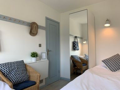 A bedroom with a bed a wicker chair with cushions a wardrobe with a mirror a door and a wall-mounted light at Little Beach in Seaview