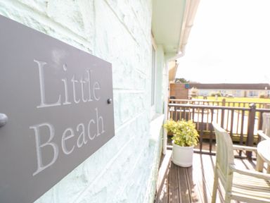 A sign reading Little Beach mounted on a light textured wall next to a wooden deck with a potted plant and outdoor furniture at Little Beach in Seaview